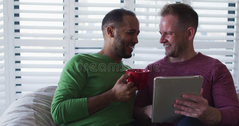 Casal homossexual multiétnico sentado no sofá um usando tablet