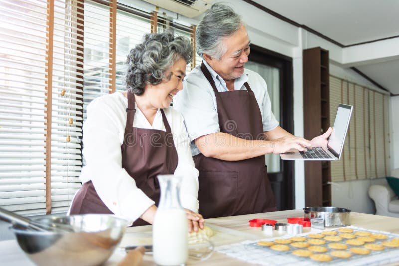 Happy Asian Elderly couple in brown aprons using laptop computer for learning online cookies class while preparing dough together. Grandfather and Grandmother having fun in a kitchen. Happy Asian Elderly couple in brown aprons using laptop computer for learning online cookies class while preparing dough together. Grandfather and Grandmother having fun in a kitchen