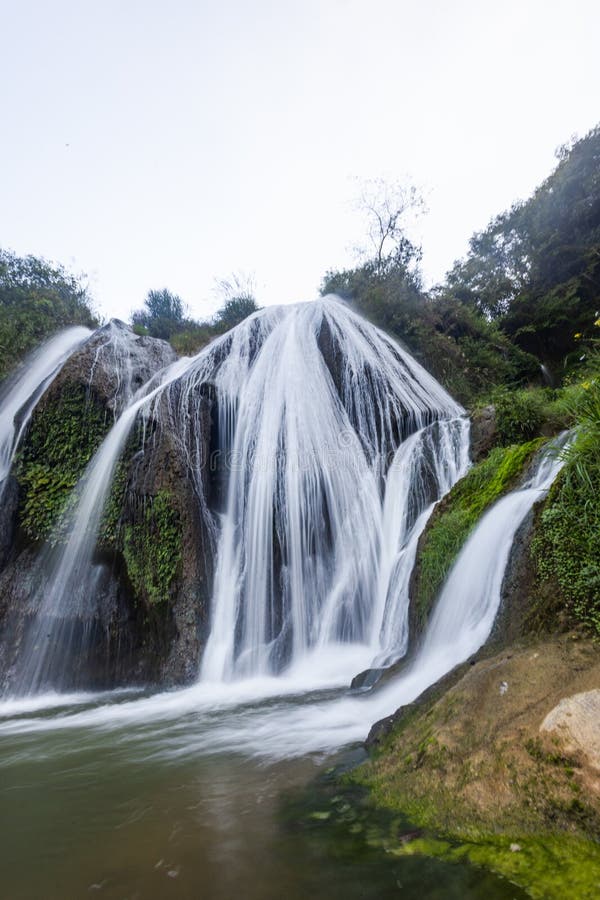 A cascading waterfall flows down over rocks, surrounded by lush greenery. The water is clear, creating a misty effect as it tumbles down. The surrounding vegetation includes moss and various plants, contributing to the dense, forested environment. The waterfall pools at the base, forming a small stream. The scene creates a sense of tranquility and natural beauty in a mountainous and forested setting. Trees and bushes frame the waterfall, enhancing the serene landscape. Fountain waterfall clear stock images, royalty-free photos and pictures