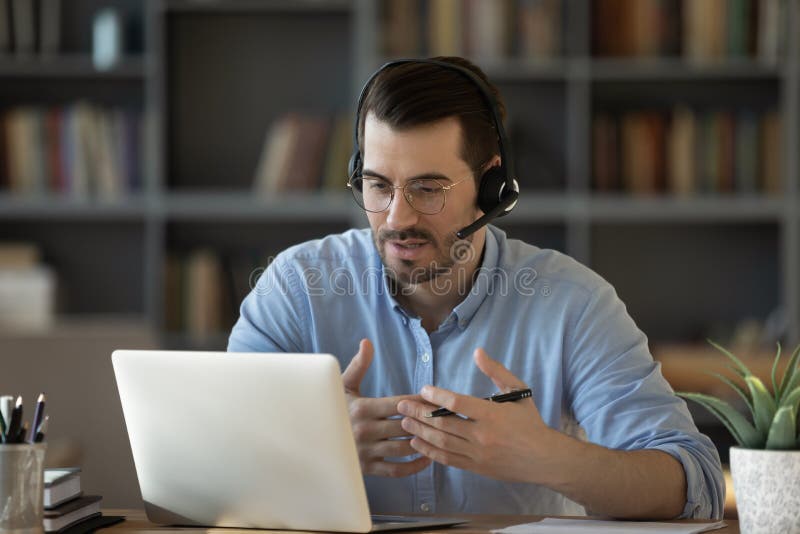 Confident man teacher coach wearing headset speaking, holding online lesson, focused student wearing glasses looking at laptop screen, studying, watching webinar training, listening to lecture. Confident man teacher coach wearing headset speaking, holding online lesson, focused student wearing glasses looking at laptop screen, studying, watching webinar training, listening to lecture