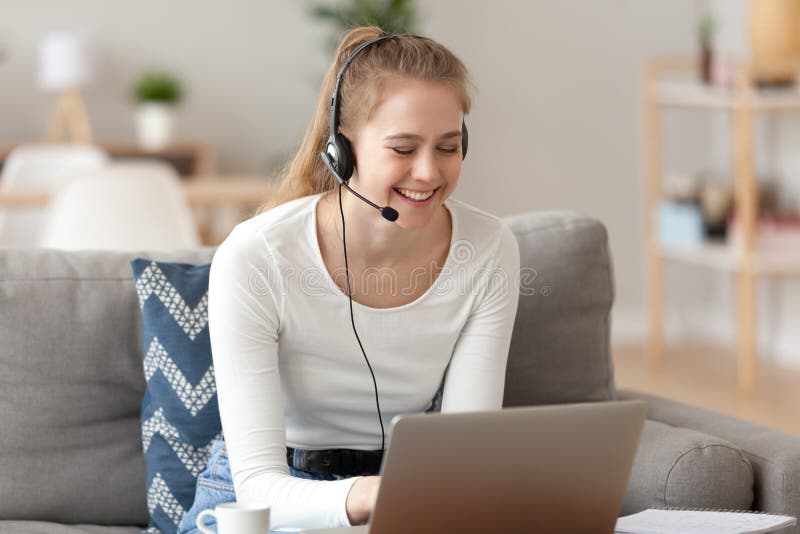 Smiling woman wearing headset with microphone, using laptop at home, sitting on sofa in living room, looking at screen, making video call, working support operator, translator, watching webinar. Smiling woman wearing headset with microphone, using laptop at home, sitting on sofa in living room, looking at screen, making video call, working support operator, translator, watching webinar