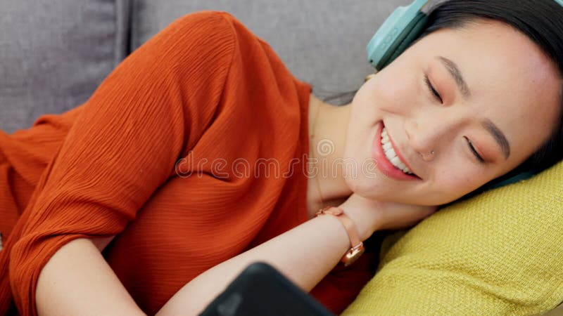 Casque de téléphone et femme streaming film tout en se relaxant sur le canapé dans sa maison moderne. rire heureux et fille asiati banque de vidéos