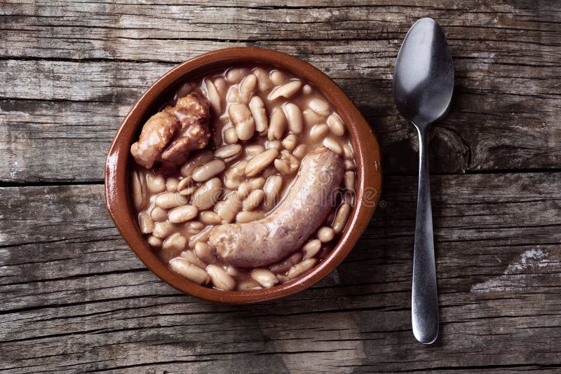 High angle view of an earthenware bowl with a cassoulet de Castelnaudary, a typical bean stew from Occitanie, in France, on a rustic wooden table. High angle view of an earthenware bowl with a cassoulet de Castelnaudary, a typical bean stew from Occitanie, in France, on a rustic wooden table