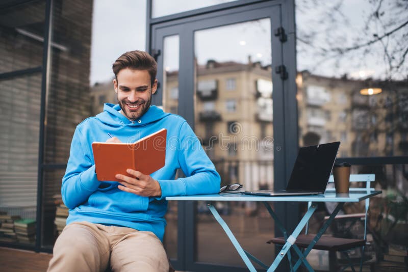Cheerful male writing education information while e learning in sidewalk cafe with good WIFI connection for web research on netbook, Caucaisan freelancer with laptop making notes in textbook. Caucaisan man stock images, royalty-free photos and pictures