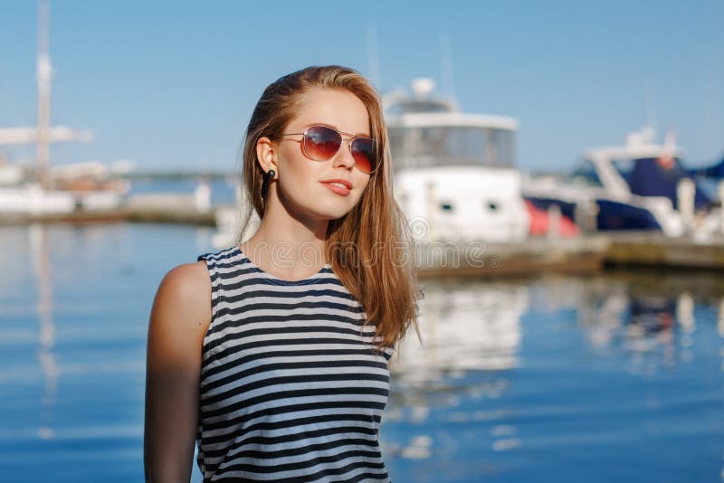 Caucasian blonde woman with tanned skin striped t-shirt and blue jeans by seashore lakeshore, with yachts boats on background