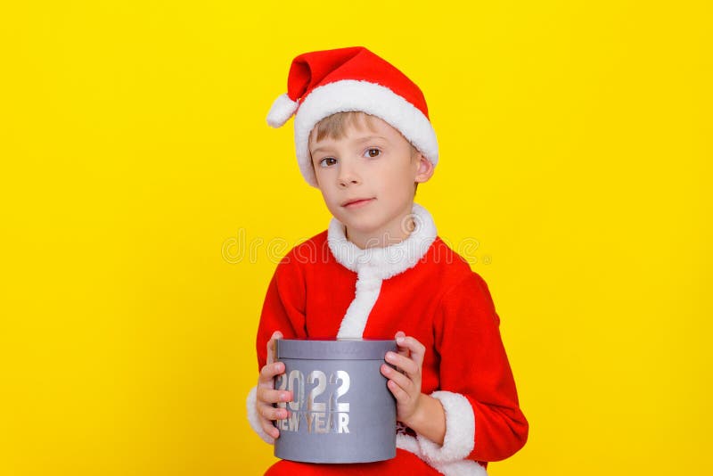 Caucasian cute child dressed as Santa Claus is holding a round gift box with the inscription New Year 2022