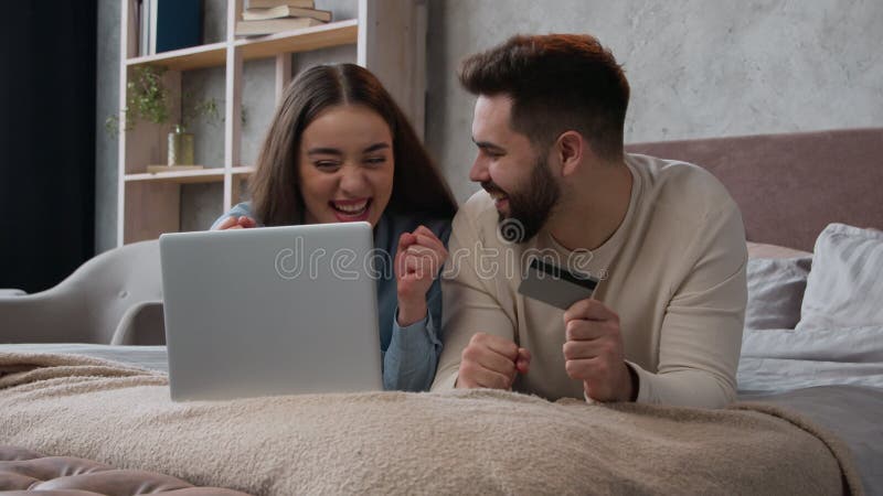 Caucasian happy couple woman and man relax on bed with laptop shopping online use bank credit card pay order buying in