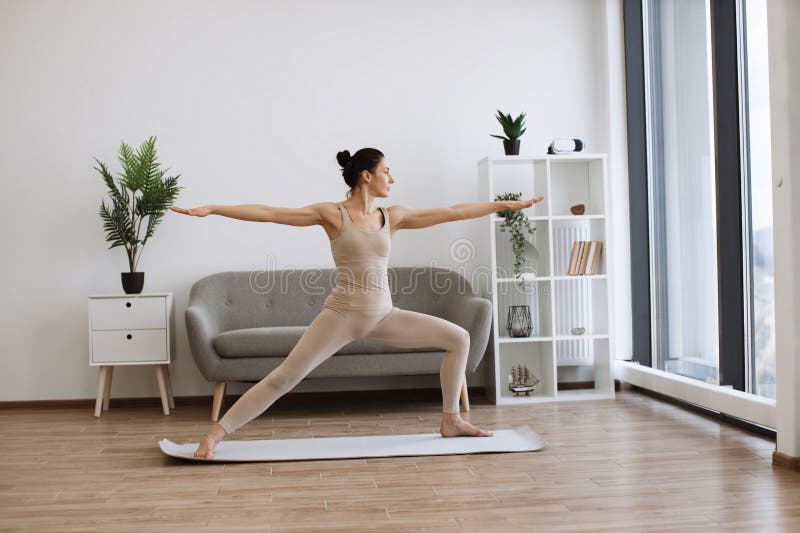 Caucasian woman spread her legs wide and pulls one knee forward, straightening arms at shoulder width in different directions. Mature brunette practising Warrior Two Pose during yoga exercise at home. Caucasian woman spread her legs wide and pulls one knee forward, straightening arms at shoulder width in different directions. Mature brunette practising Warrior Two Pose during yoga exercise at home.