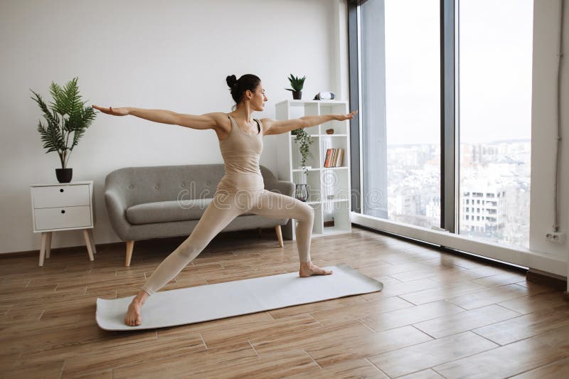 Caucasian woman spread her legs wide and pulls one knee forward, straightening arms at shoulder width in different directions. Mature brunette practising Warrior Two Pose during yoga exercise at home. Caucasian woman spread her legs wide and pulls one knee forward, straightening arms at shoulder width in different directions. Mature brunette practising Warrior Two Pose during yoga exercise at home.