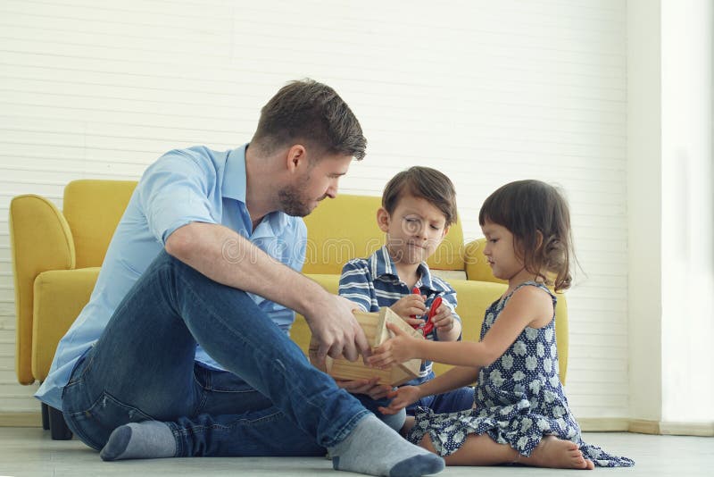 A Caucasian young father sitting on warm floor playing wooden house blocks with happy little son and daughter