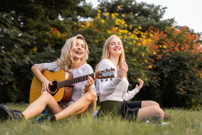 caucasian young women sitting in the park outdoor and playing a guitar sing a song together with happiness