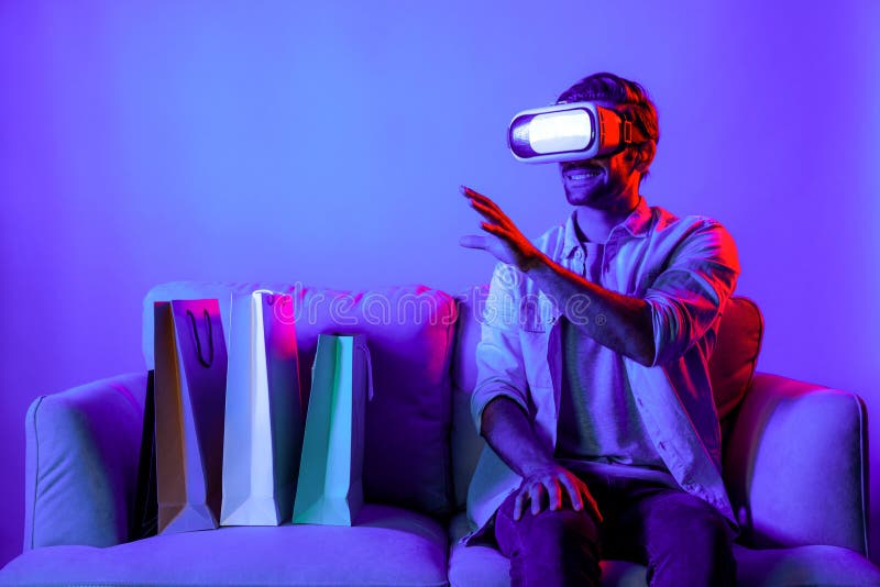 Caucasian man with vr glass posing pointing finger while sitting at sofa with shopping bags. Person selecting and doing shopping online by using VR technology and virtual goggle at home. Deviation. Caucasian man with vr glass posing pointing finger while sitting at sofa with shopping bags. Person selecting and doing shopping online by using VR technology and virtual goggle at home. Deviation.