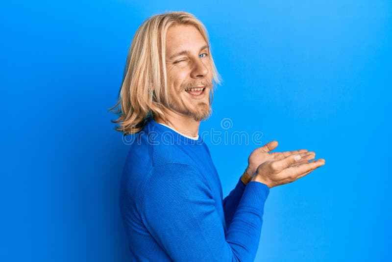 Caucasien jeune homme avec les cheveux longs présentant avec des paumes ouvertes tenant quelque chose de clin d'oeil à la caméra a photo stock