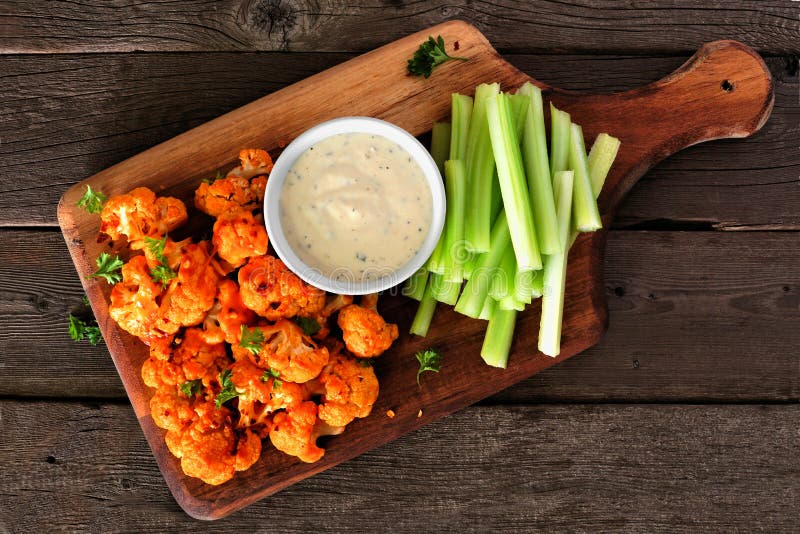 Healthy vegan cauliflower buffalo wings with celery and ranch dip, top view on a wood paddle board
