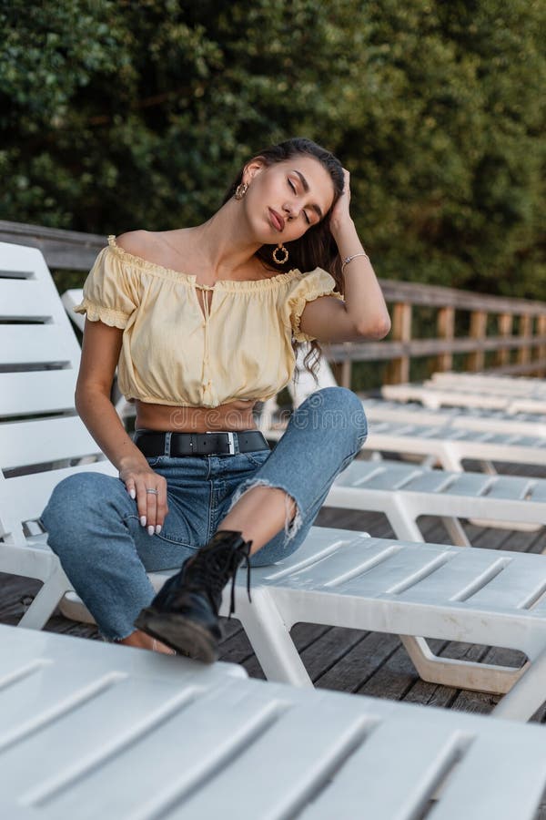 Całkiem szczęśliwa hipster kobieta w modzie niebieskie dżinsy i żółty wierzchołek z czarnymi butami spoczywają na lodzie na plaży fotografia stock