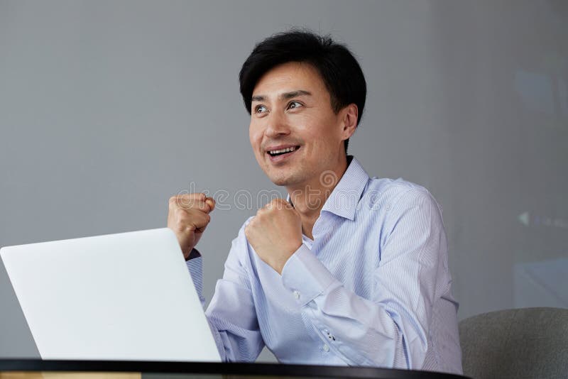 Celebrating success. Excited young Asian businessman arms up while working at office with laptop. stock images Celebrating success. Excited young Asian businessman arms up while working at office with laptop. stock images