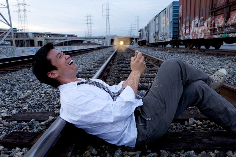 An Asian man sitting on the train tracks making a call.