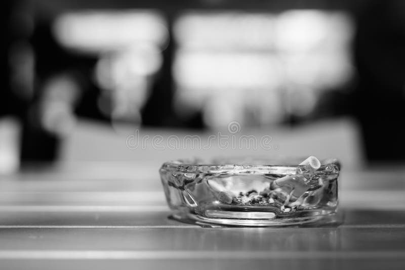 Ashtray with cigarettes on wooden table in restaurant with out of focus background and bokeh effect in black and white. Ashtray with cigarettes on wooden table in restaurant with out of focus background and bokeh effect in black and white