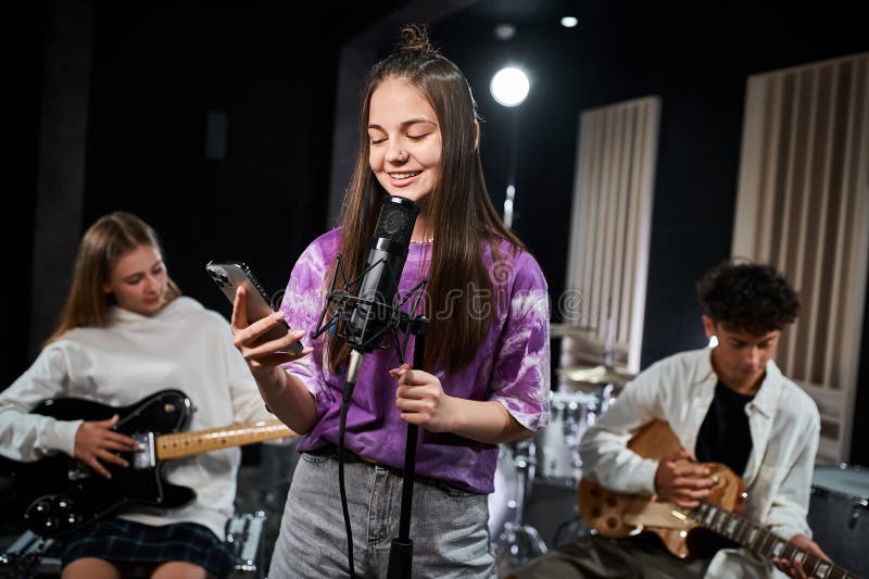 focus on cheerful teenage girl singing and looking at her phone next to her blurred guitarists, stock photo. focus on cheerful teenage girl singing and looking at her phone next to her blurred guitarists, stock photo