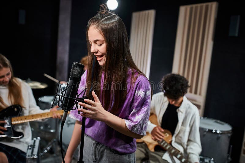 focus on cute teenage girl in vivid attire singing next to her blurred band members in studio, stock photo. focus on cute teenage girl in vivid attire singing next to her blurred band members in studio, stock photo