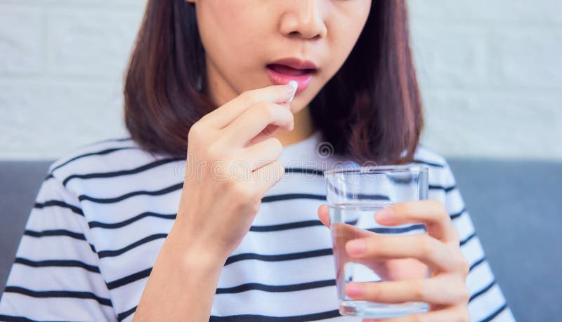 Cerca de una mujer tomando pastillas blancas en la boca y bebiendo agua en vidrio en el sofá de la casa, se siente como enferma C