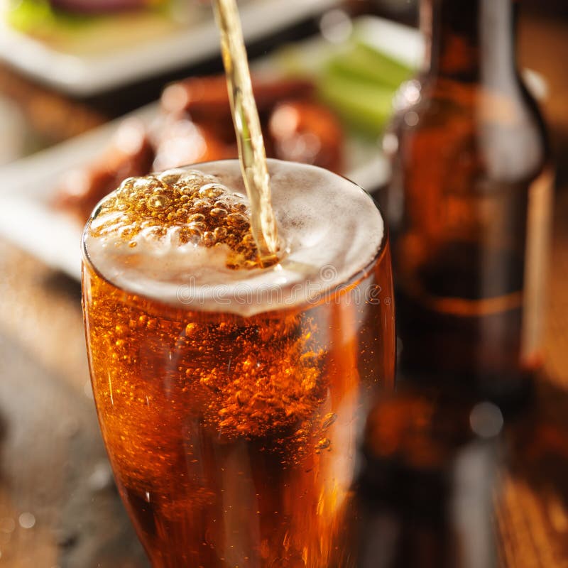 Pouring beer into glass with bbq chicken wings in background. Pouring beer into glass with bbq chicken wings in background