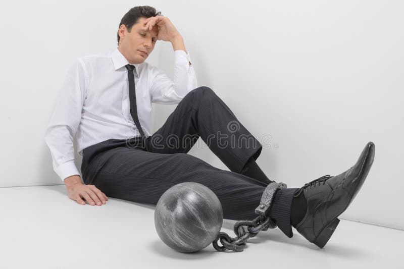 Chained businessman. Full length of depressed businessman sitting on the floor with shackles chained to his legs