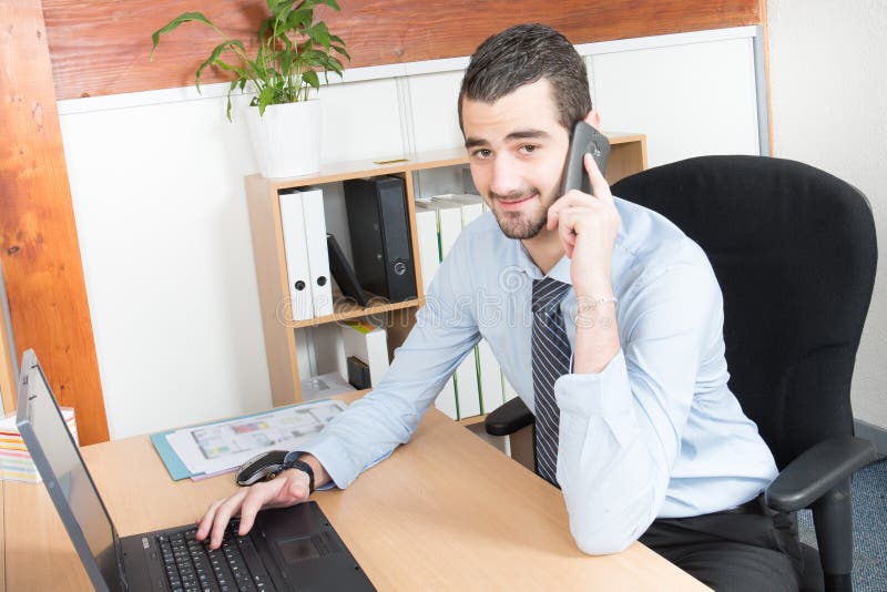 chamando o homem de negócios do telefone que senta-se no escritório e em usar seu portátil ao trabalhar no projeto novo foto de stock royalty free chamando o homem de negócios do telefone que senta-se no escritório e em usar seu portátil ao trabalhar no projeto novo foto de stock royalty free