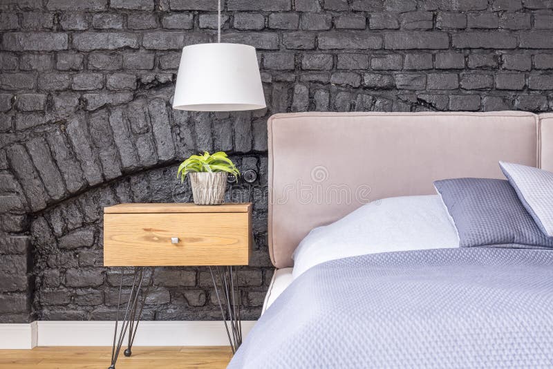 Small plant in pot on wooden bedside cabinet under white lamp in bedroom with brick wall. Small plant in pot on wooden bedside cabinet under white lamp in bedroom with brick wall