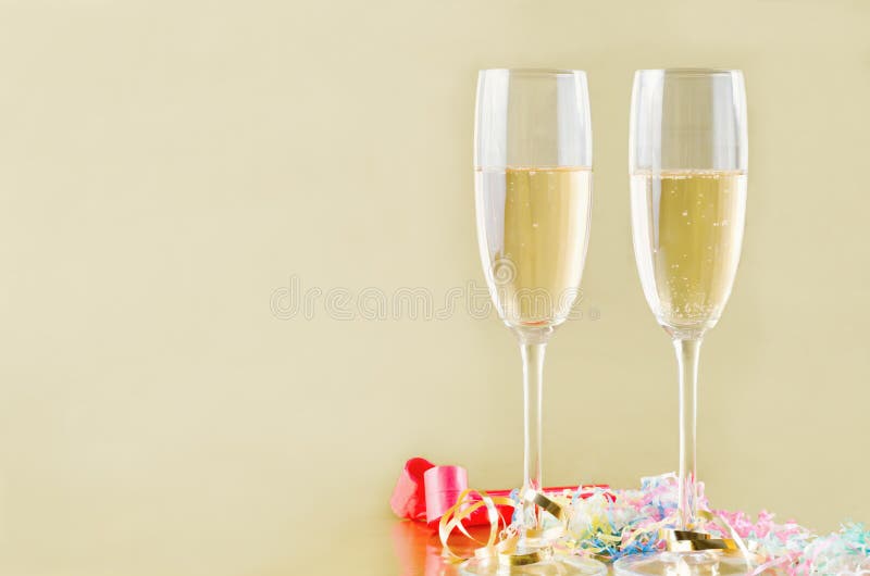 Two fluted champagne glasses with bubbles rising on a gold background with popper streamers and party horn blowers on the surface below. Copy space to the left. Two fluted champagne glasses with bubbles rising on a gold background with popper streamers and party horn blowers on the surface below. Copy space to the left.