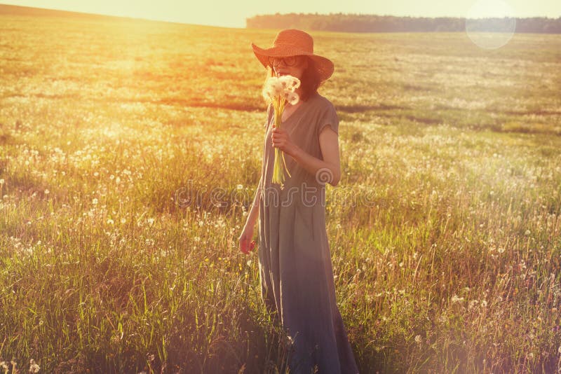 Chapeau de paille de port de femme et longue robe de toile appr?ciant la nature, tenant le bouquet de beaux pissenlits frais, jou image stock Chapeau de paille de port de femme et longue robe de toile appr?ciant la nature, tenant le bouquet de beaux pissenlits frais, jou image stock