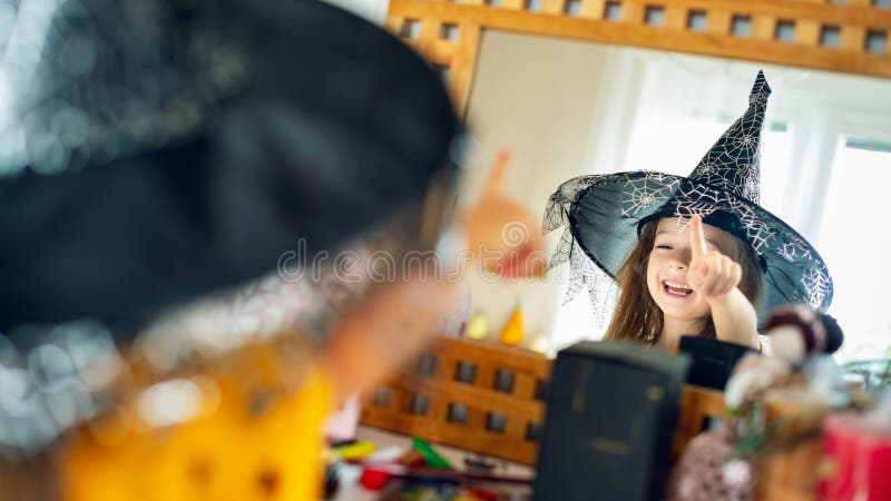 Adorable young girl wearing witch hat looking at herself in the mirror, pointing with finger and laughing. Happy child in halloween costume looking in the mirror. Adorable young girl wearing witch hat looking at herself in the mirror, pointing with finger and laughing. Happy child in halloween costume looking in the mirror.