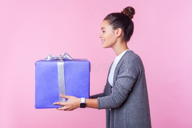 Charity donation. Side view of generous teenage brunette girl giving gift box with kind smile, pink background