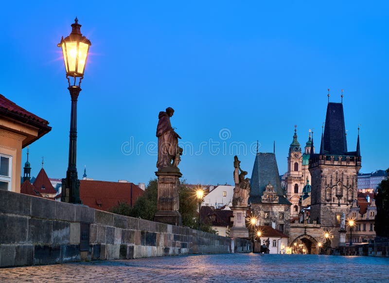 Charles Bridge at dawn, silhouette of Bridge Tower and saint sculptures with lit street light lamp in Prague, Czech Republic