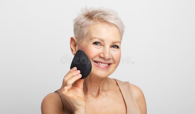 Charmante vieille femme avec cheveux courts utilisation éponge enlever maquillage sur fond gris clair. photo libre de droits