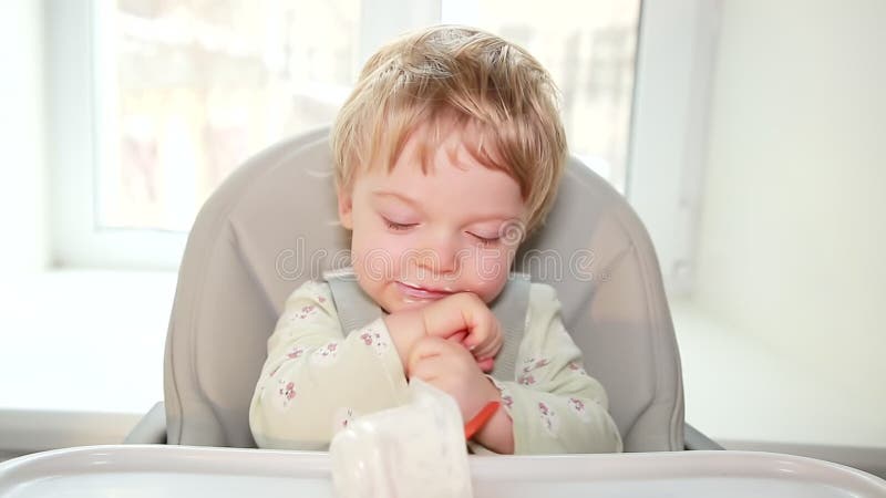 A charming boy has eaten a baby curd from a jar and is sitting contentedly in his chair