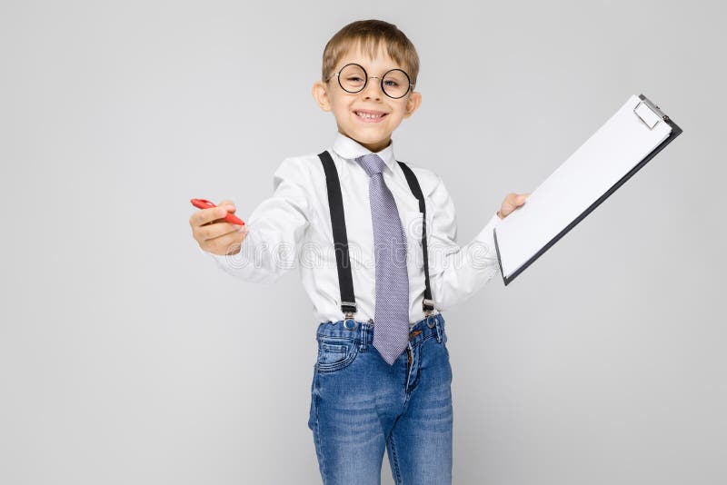 A charming boy in a white shirt, suspenders, a tie and light jeans stands on a gray background. The boy holds a pen and