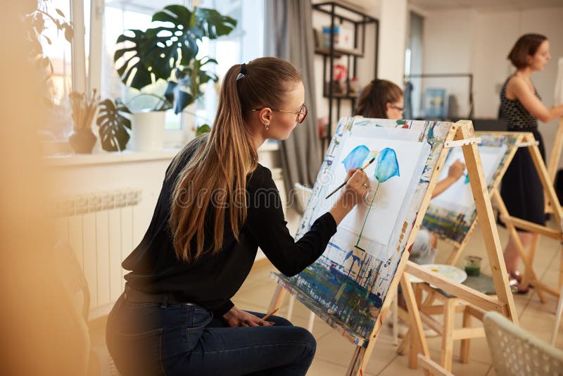 Charming fair-haired girl in glasses dressed in black blouse and jeans sits at the easel and paints a picture in the art stock images Charming fair-haired girl in glasses dressed in black blouse and jeans sits at the easel and paints a picture in the art stock images
