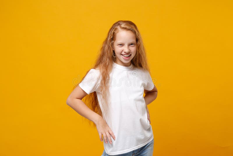 Charming little ginger kid girl 12-13 years old in white t-shirt isolated on yellow background children portrait. Childhood lifestyle concept. Mock up copy space. Standing with arms akimbo on waist