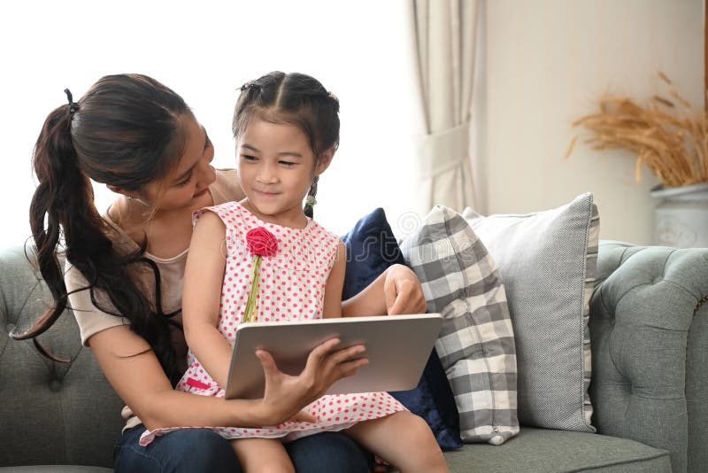 Charming little girl and her beautiful young mom are using a digital tablet on couch. stock photos