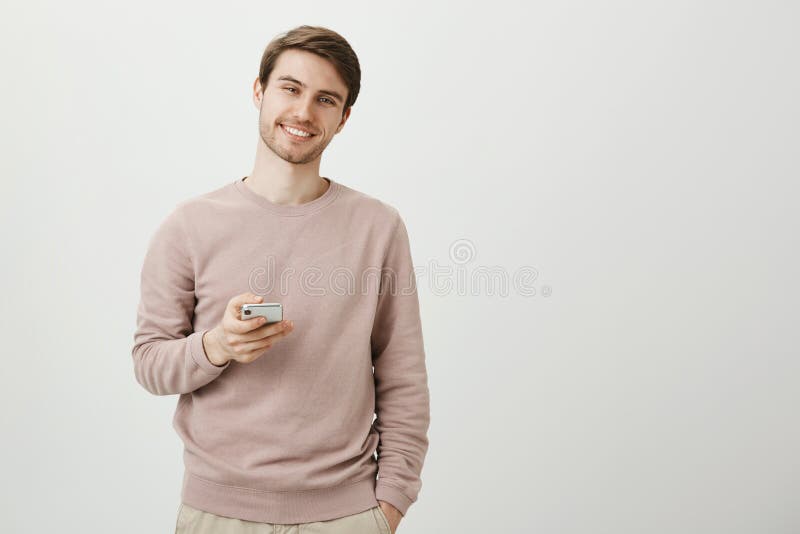 Charming pleasant young unshaven man standing with cute smile, holding smartphone and looking at camera while being over stock photos Charming pleasant young unshaven man standing with cute smile, holding smartphone and looking at camera while being over stock photos