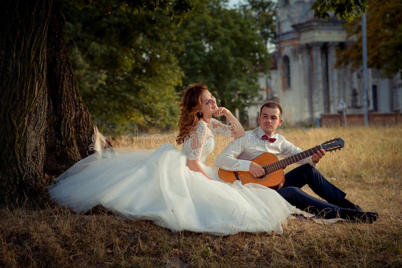 Charming smiling bride is listening to the elegant groom playing the guitar while sitting on the grass under the tree.