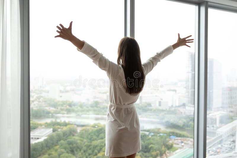 That is my world. Charming young woman in bathrobe standing at large panoramic window wall stretching hands to sides opening embraces to life enjoying splendid city panorama meeting new day, back view. That is my world. Charming young woman in bathrobe standing at large panoramic window wall stretching hands to sides opening embraces to life enjoying splendid city panorama meeting new day, back view