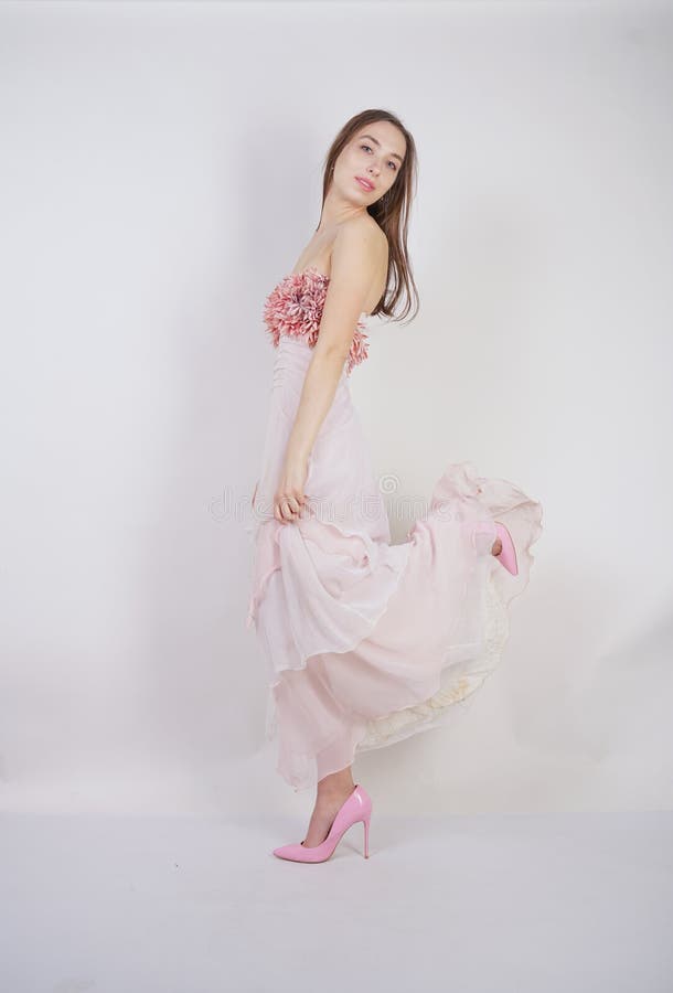 A charming young caucasian girl stands in a pink long prom dress with flower petals on her chest and poses on a white background i stock photo A charming young caucasian girl stands in a pink long prom dress with flower petals on her chest and poses on a white background i stock photo