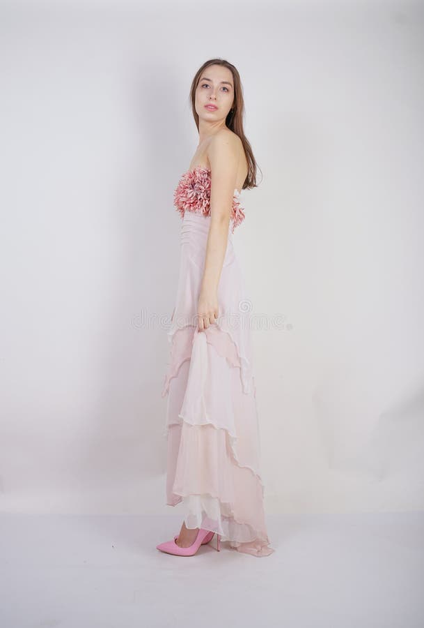 A charming young caucasian girl stands in a pink long prom dress with flower petals on her chest and poses on a white background i stock photo A charming young caucasian girl stands in a pink long prom dress with flower petals on her chest and poses on a white background i stock photo