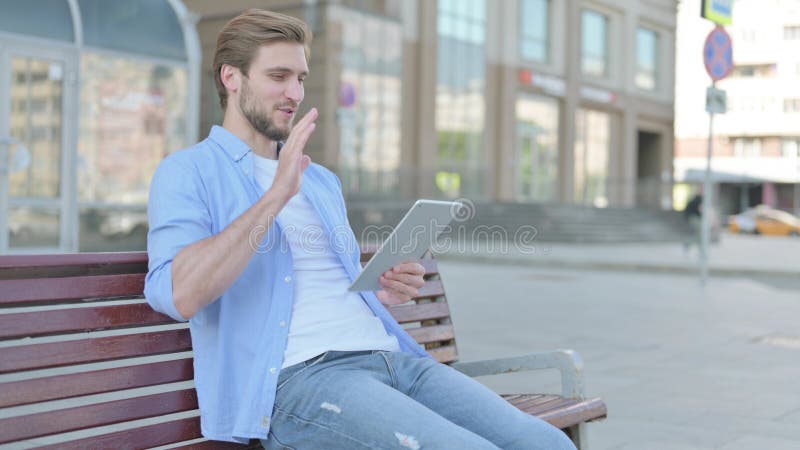 Chat de video en línea en tablet por un hombre sentado al aire libre en el banco fotografía de archivo libre de regalías