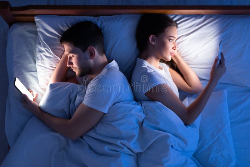 Cheating Concept. Husband And Wife Texting On Phones Lying Back-To-Back In Bed In Bedroom. Top View, Low Light. Cheating Concept. Husband And Wife Texting On Phones Lying Back-To-Back In Bed In Bedroom. Top View, Low Light