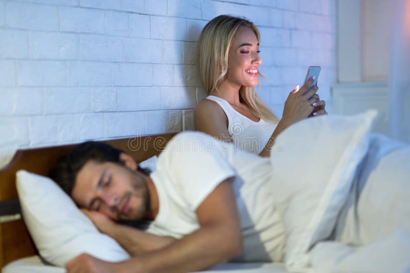 Cheating Wife Chatting On Cellphone While Her Husband Sleeping In Bedroom At Night. Low Light, Selective Focus. Cheating Wife Chatting On Cellphone While Her Husband Sleeping In Bedroom At Night. Low Light, Selective Focus