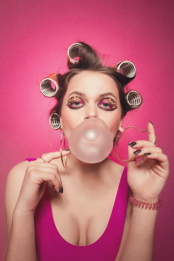 Cheeky girl with bubble gum posing on pink background in body, with curlers on head. 