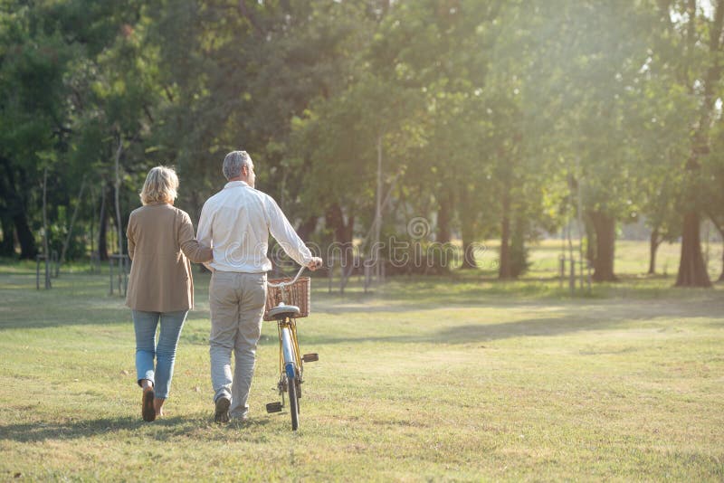 Cheerful active senior couple with bicycle walking through park together. Perfect activities for elderly people in retirement lifestyle.